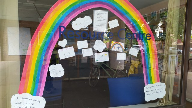 Large rainbow painted on a window with handwritten notes and drawings displayed at a resource centre.
