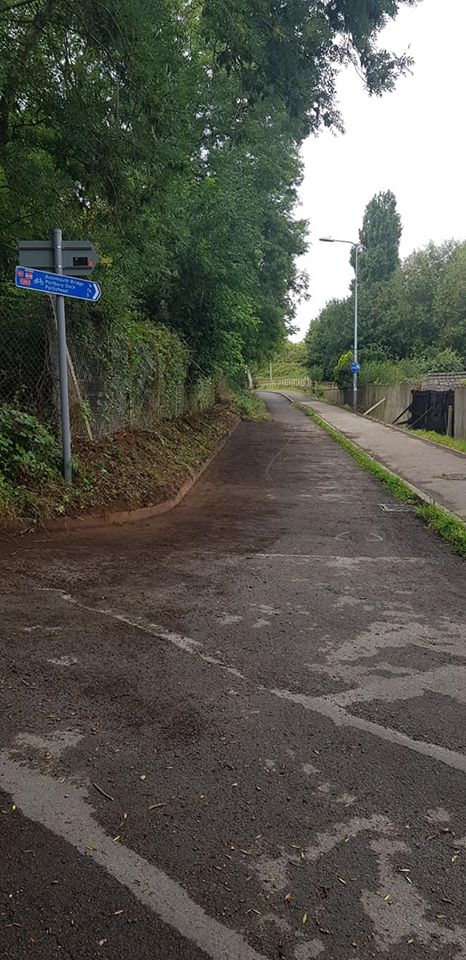 A paved path runs through trees, with a blue cycle route sign and a streetlamp along the way.