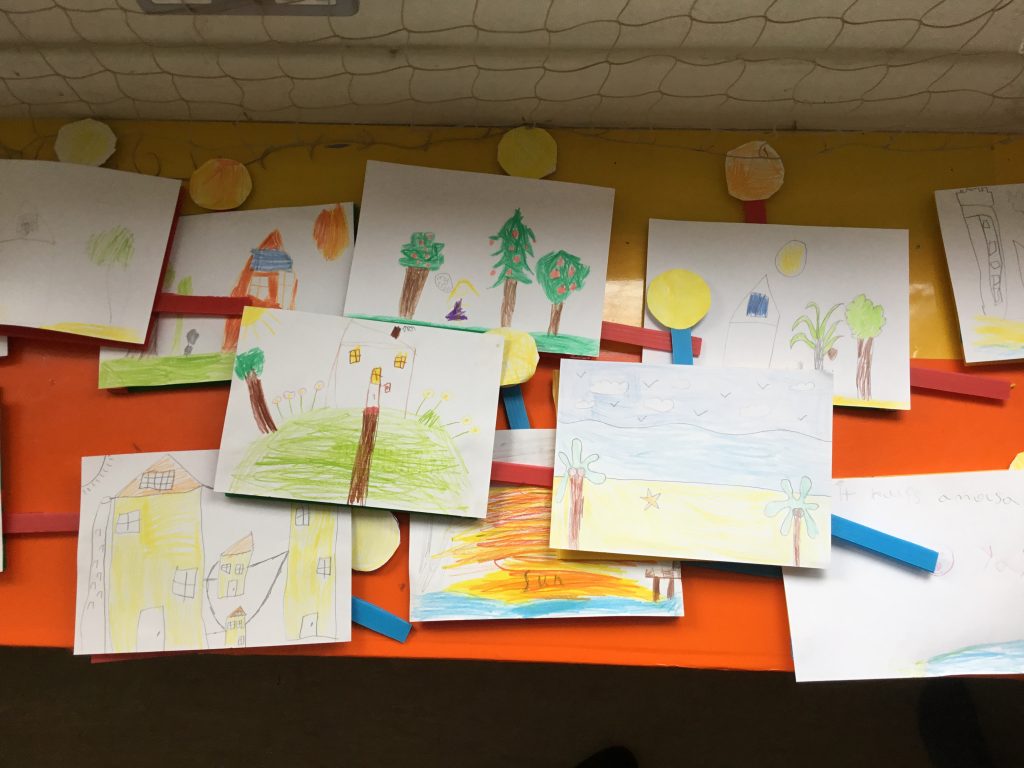 Children’s colourful drawings of houses, trees, and landscapes spread out on a table with bright sticks.