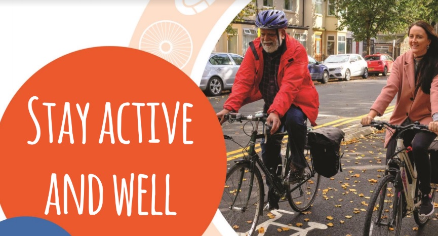 Two people wearing jackets and helmets ride bicycles on a city street with the text "Stay active and well.