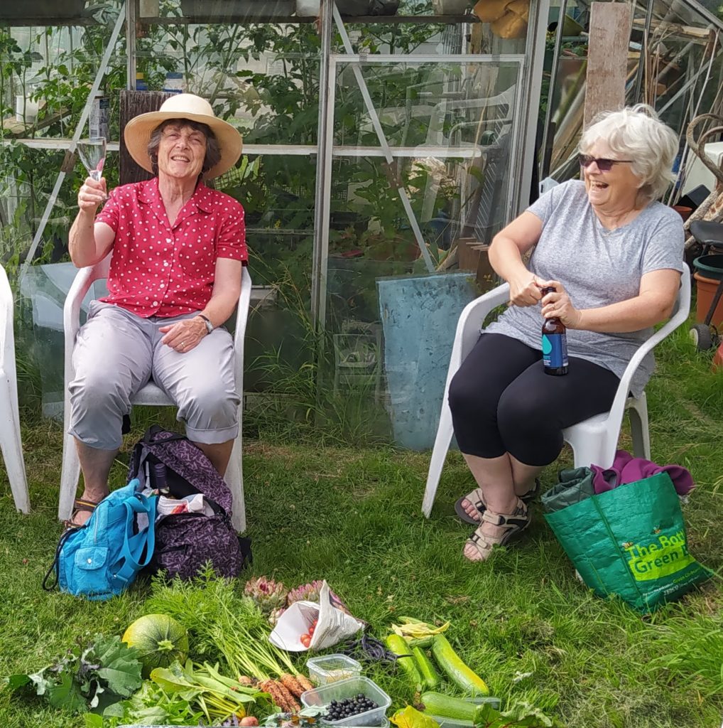 Two women sitting on chairs outdoors, smiling, with fresh vegetables and fruit laid out on the grass in front of them.