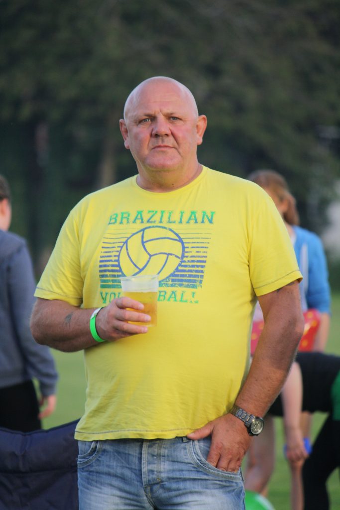 A man in a yellow "Brazilian Volleyball" shirt holds a drink whilst standing outdoors.