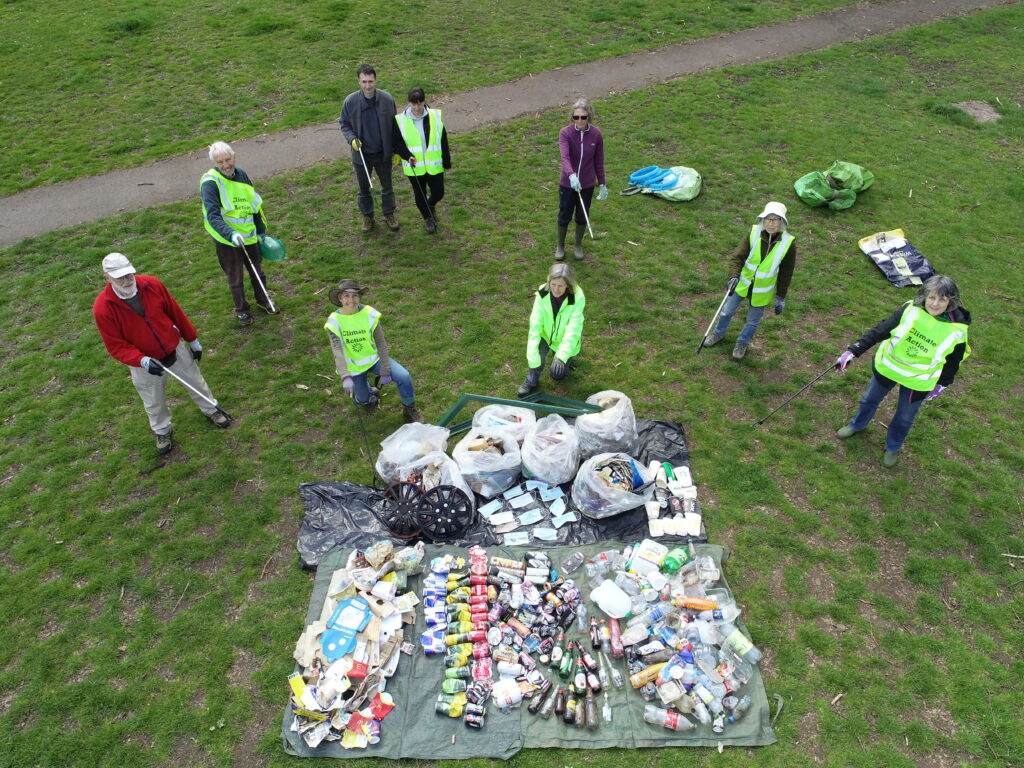 A group of people in safety vests stand around collected rubbish bags on a grassy field during a cleanup.