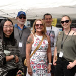 Seven smiling adults pose together under a white marquee outdoors, wearing name badges and casual clothing.