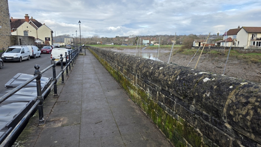A stone path runs beside a mossy wall, with houses, cars, and a cloudy sky in the background.